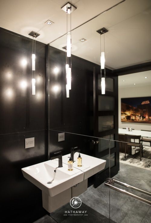 Modern bathroom with a white double wall-mount sink, black tiled wall, mirrored surface, and hanging pendant lights.