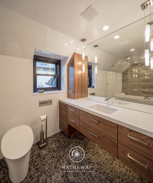 A modern bathroom with a wood vanity, white countertop, stone pebble floor, white toilet, and a large wall mirror.