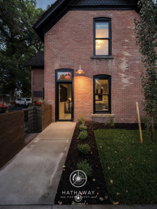 A brick house exterior at dusk with a lighted doorway, a concrete walkway, and a landscaped front garden.