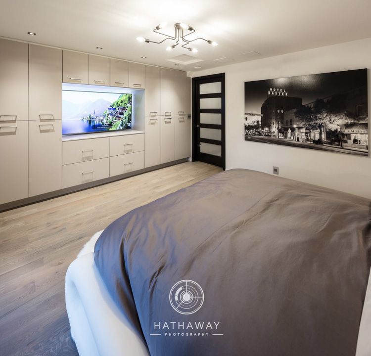A modern, minimalist bedroom with light grey walls, white built-in storage cabinets, a flat-screen TV, and a grey bed.