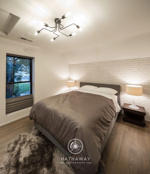 A bedroom with a grey bed, white brick wall accent, two bedside lamps, a shaggy rug, and a modern light fixture.