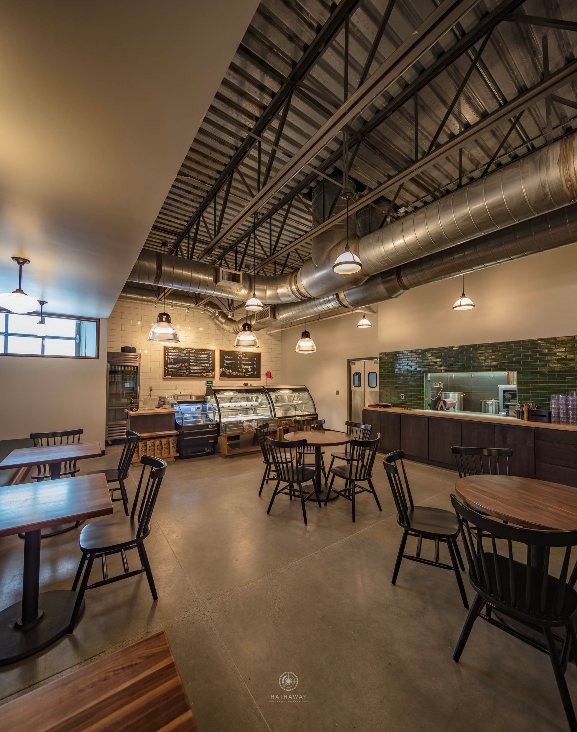 Empty tables and chairs in a cafe with industrial-style ceilings, exposed ductwork, and a food counter in the background.