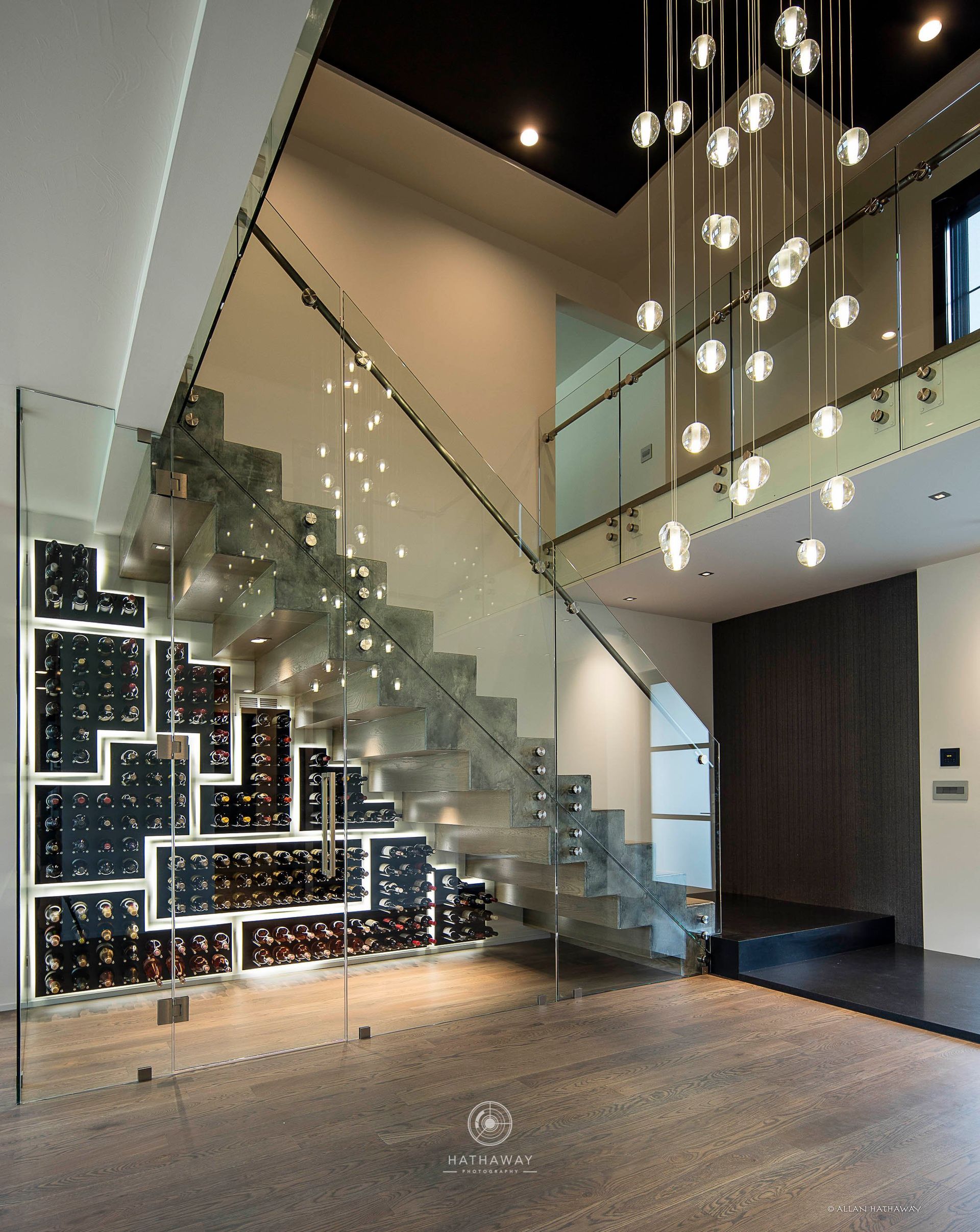 Modern interior with a glass-enclosed wine cellar beneath a staircase, illuminated by a contemporary hanging chandelier.