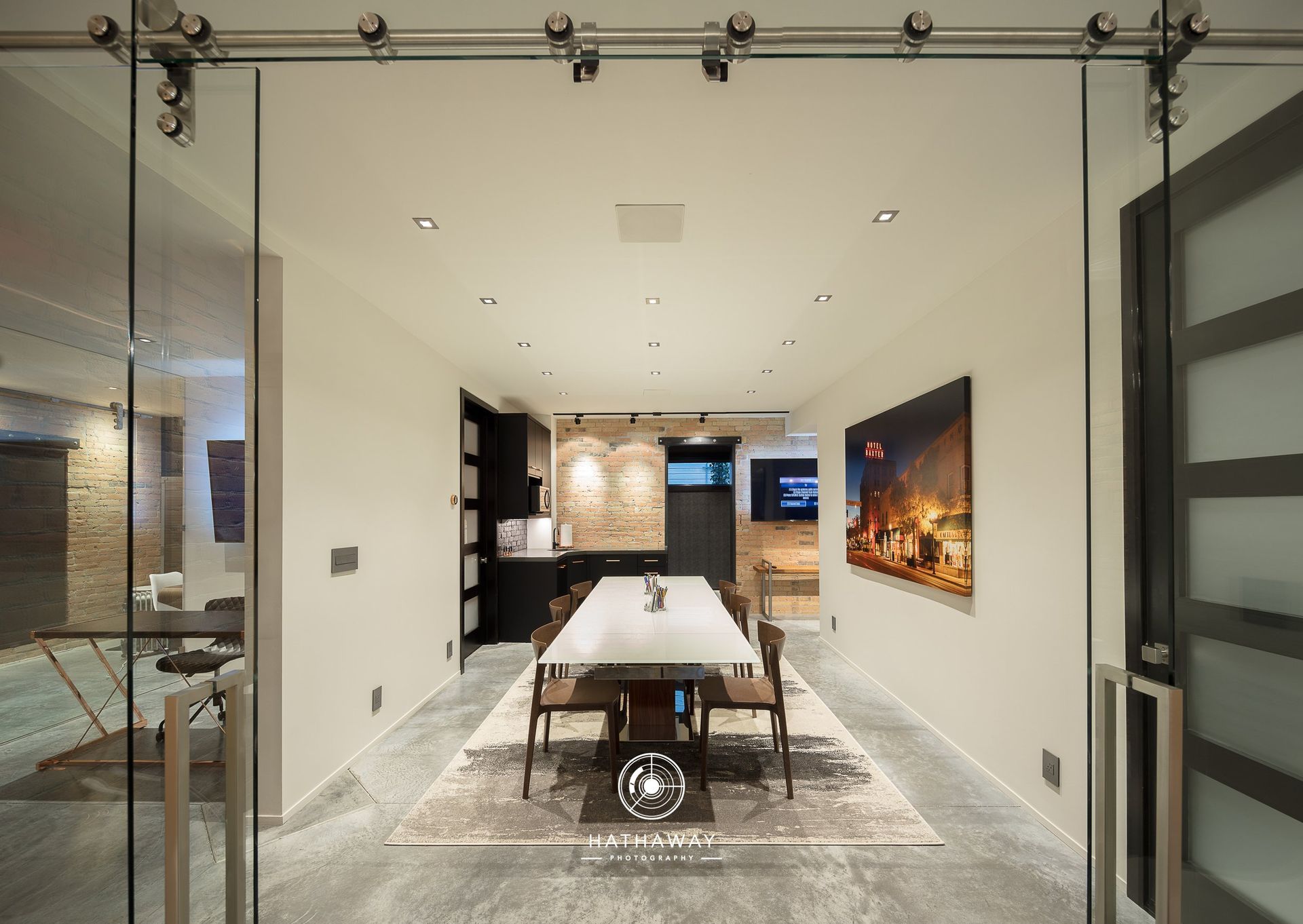A modern, neutral-toned dining room viewed through glass sliding doors, featuring a long white table and ambient lighting.