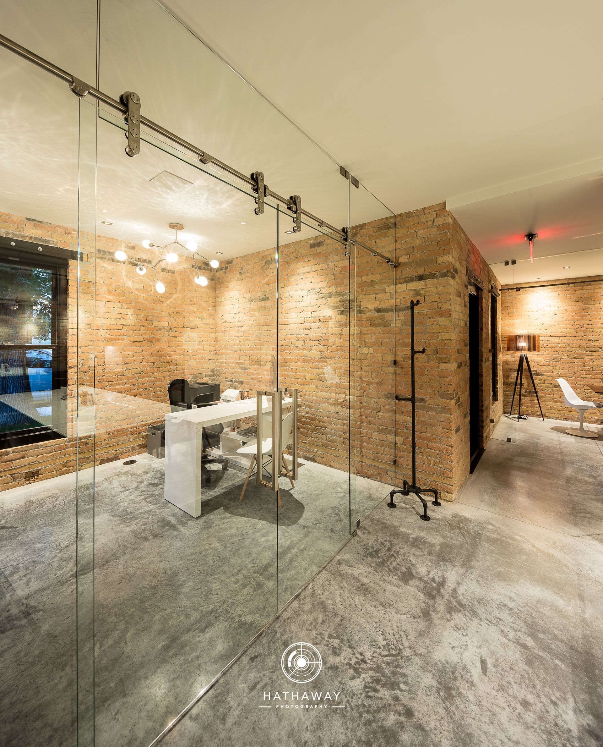Modern office space with a glass partition wall, a white desk, and exposed brick walls on a polished concrete floor.