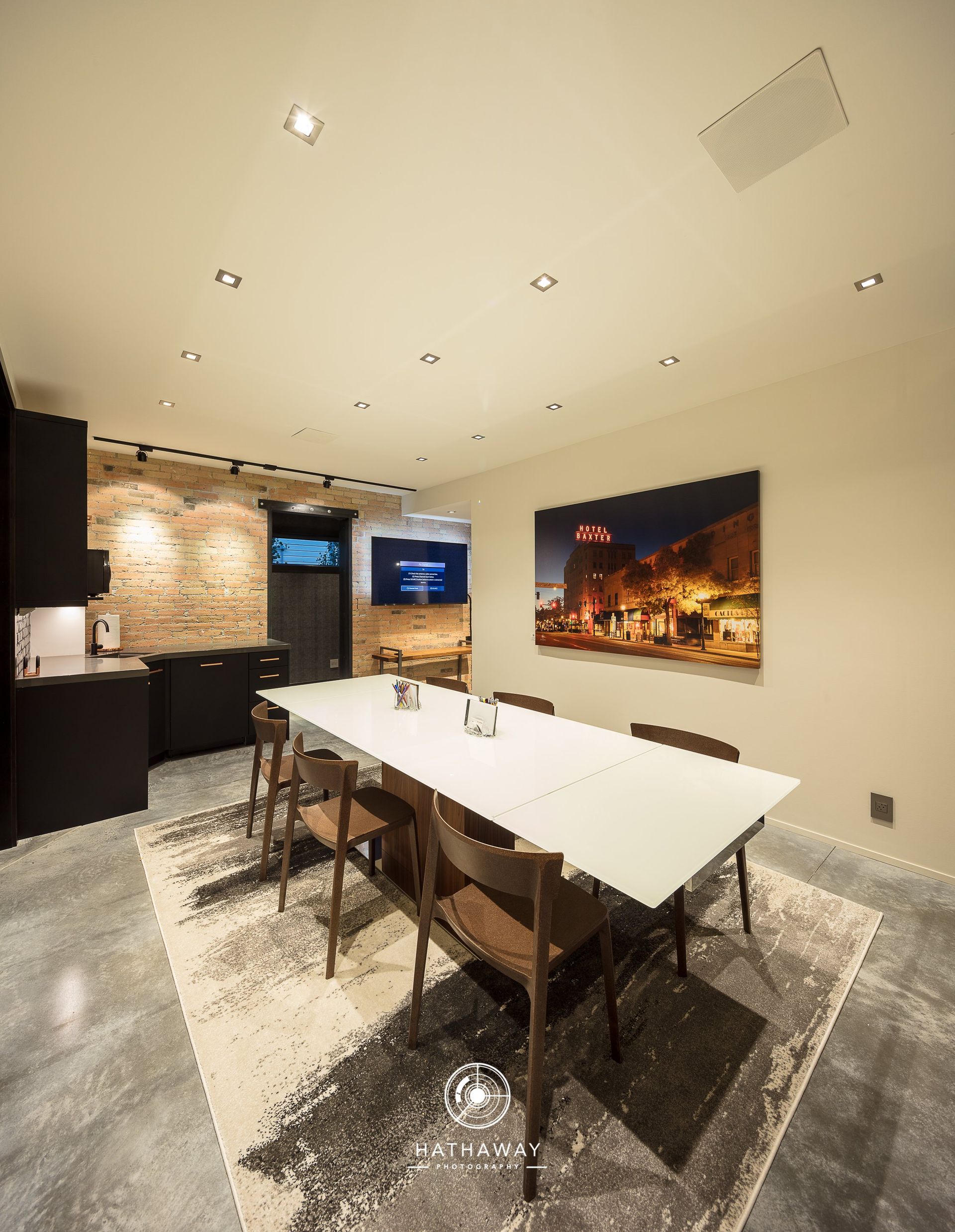 A modern dining room with a long white table, brown chairs on a textured rug, and a large landscape art piece on the wall.
