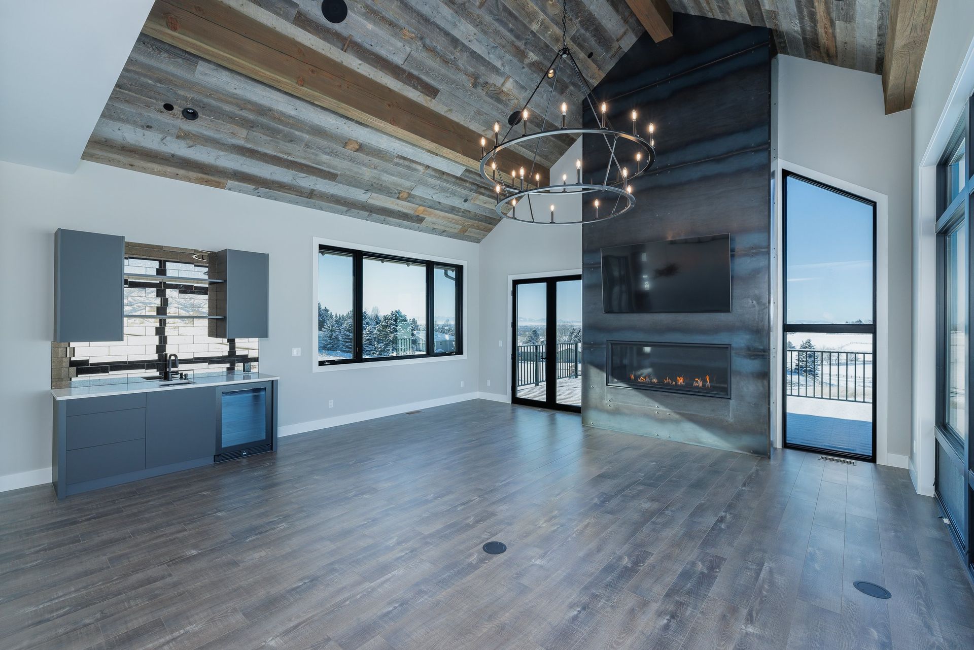 A modern living room with a vaulted wood ceiling, dark metal fireplace, large windows, and a built-in wet bar.