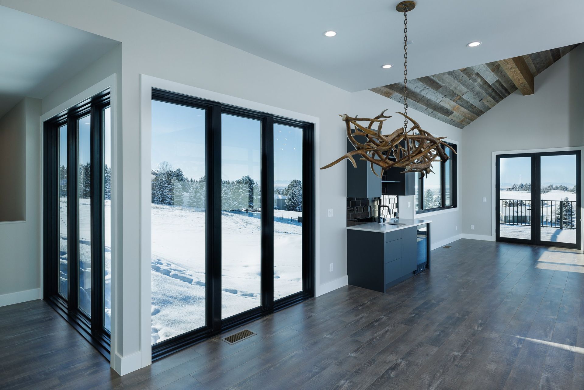 A modern living area with dark flooring, large black-framed glass doors, a rustic chandelier, and a snowy exterior view.