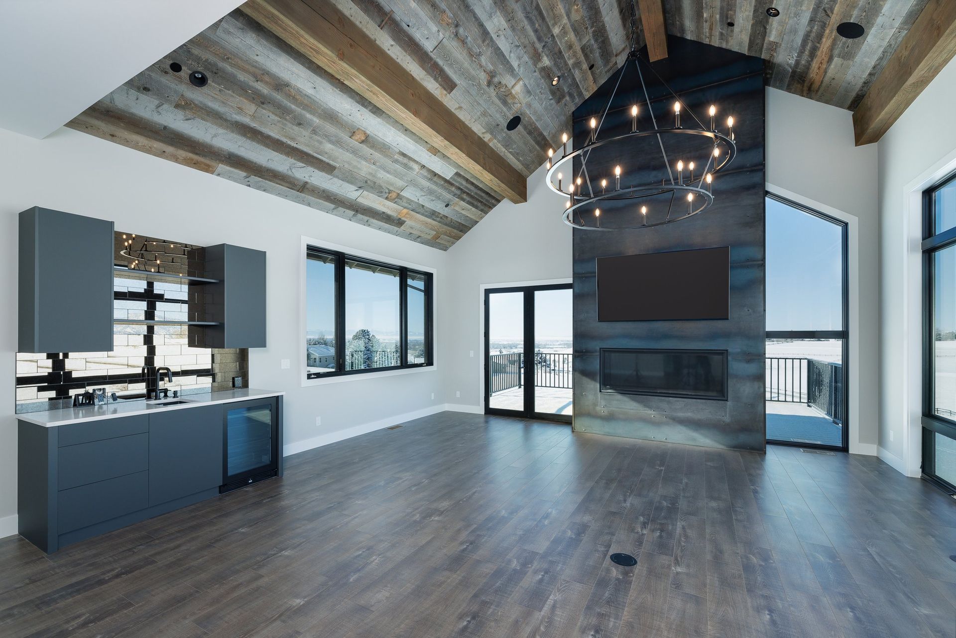 A modern living room with a vaulted wood ceiling, gray cabinets, a fireplace wall, and large windows with a view.