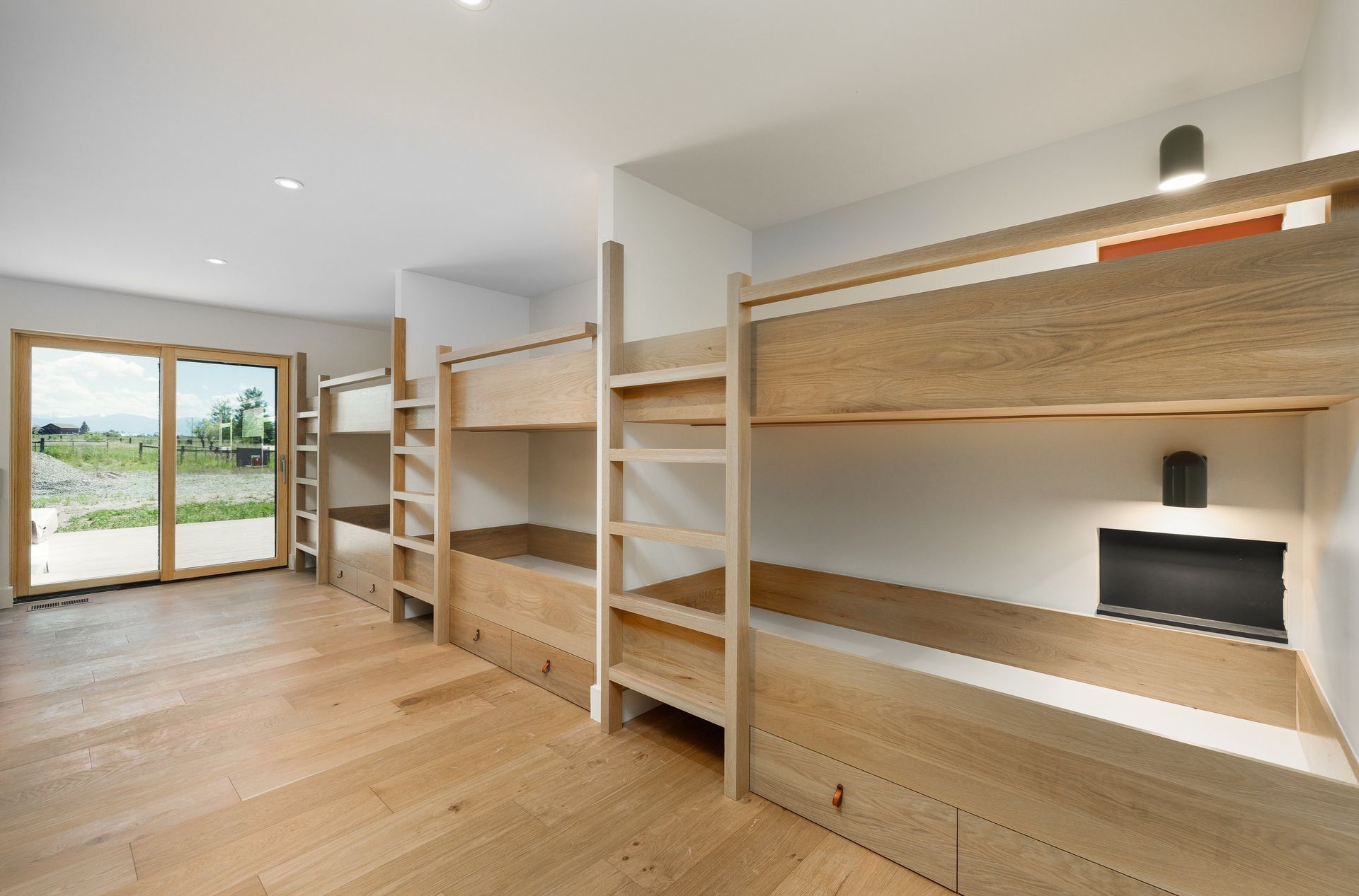A bright room with light wood floors and two sets of built-in wooden bunk beds, featuring large windows overlooking a field.