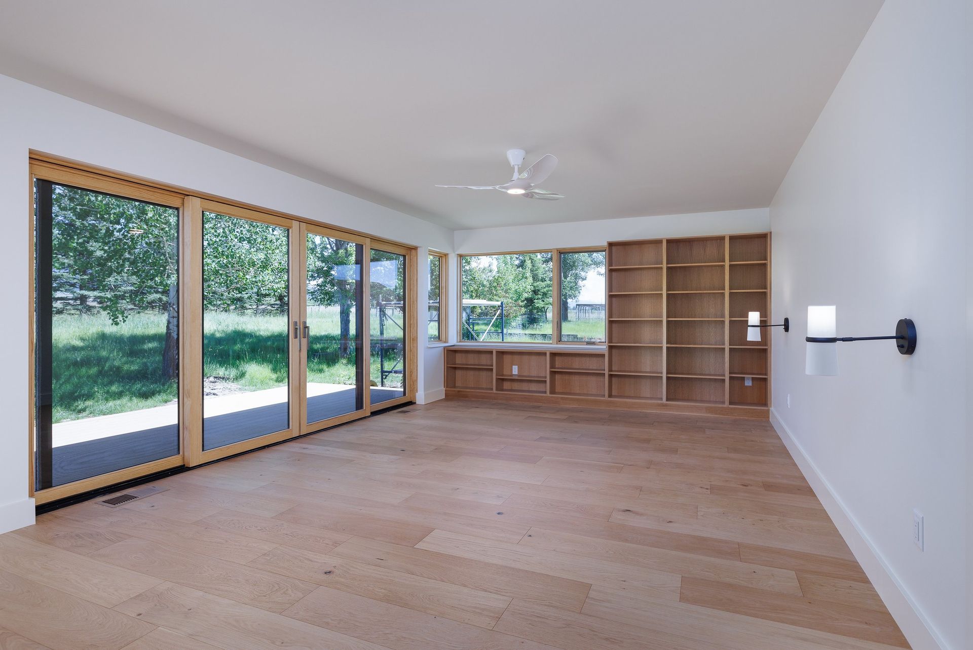 A bright room with light wood floors, a large sliding glass door, wooden bookshelves, and a ceiling fan.