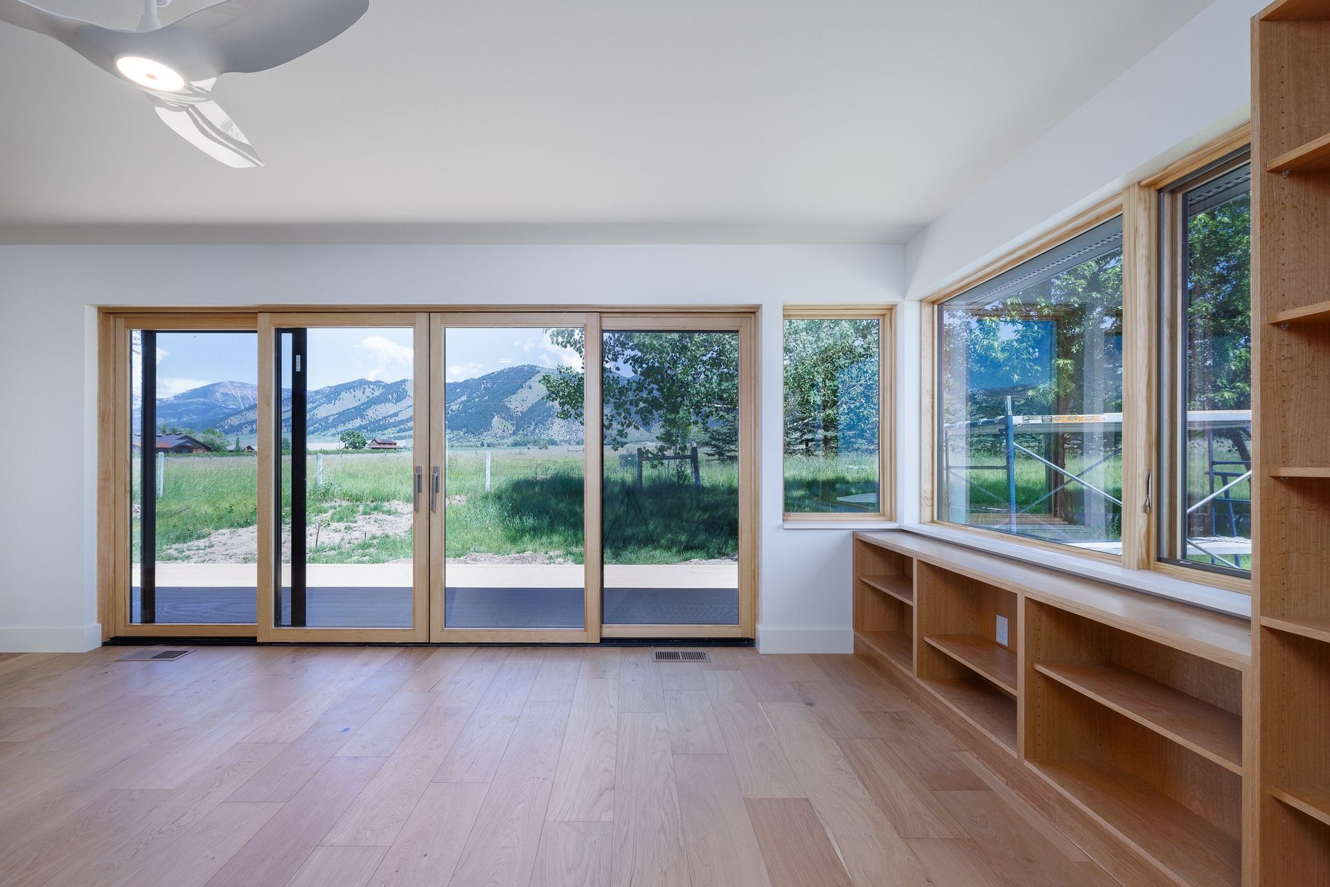 A bright, modern room featuring light wood flooring, a ceiling fan, built-in wooden bookshelves, and expansive glass doors.