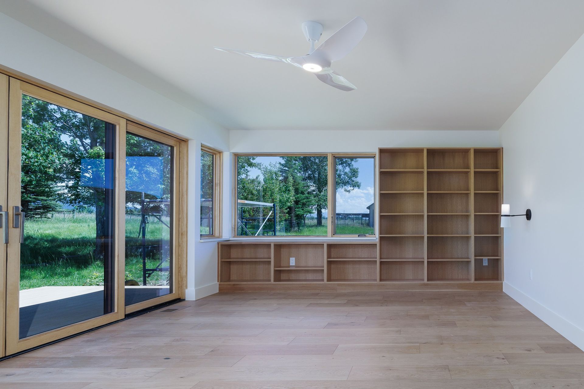 A bright, modern room with hardwood floors, a wall of built-in wooden shelving, a large window, and a glass sliding door.