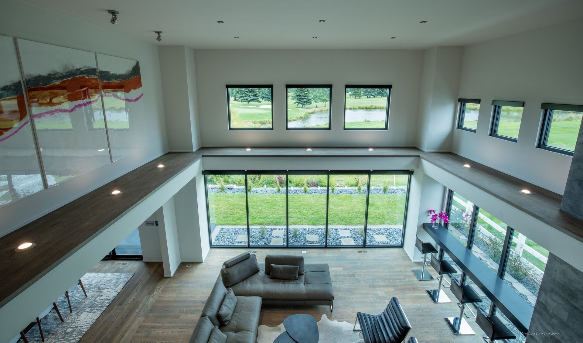 A modern, high-ceiling living room with floor-to-ceiling windows, gray sectional seating, and a sleek bar area.