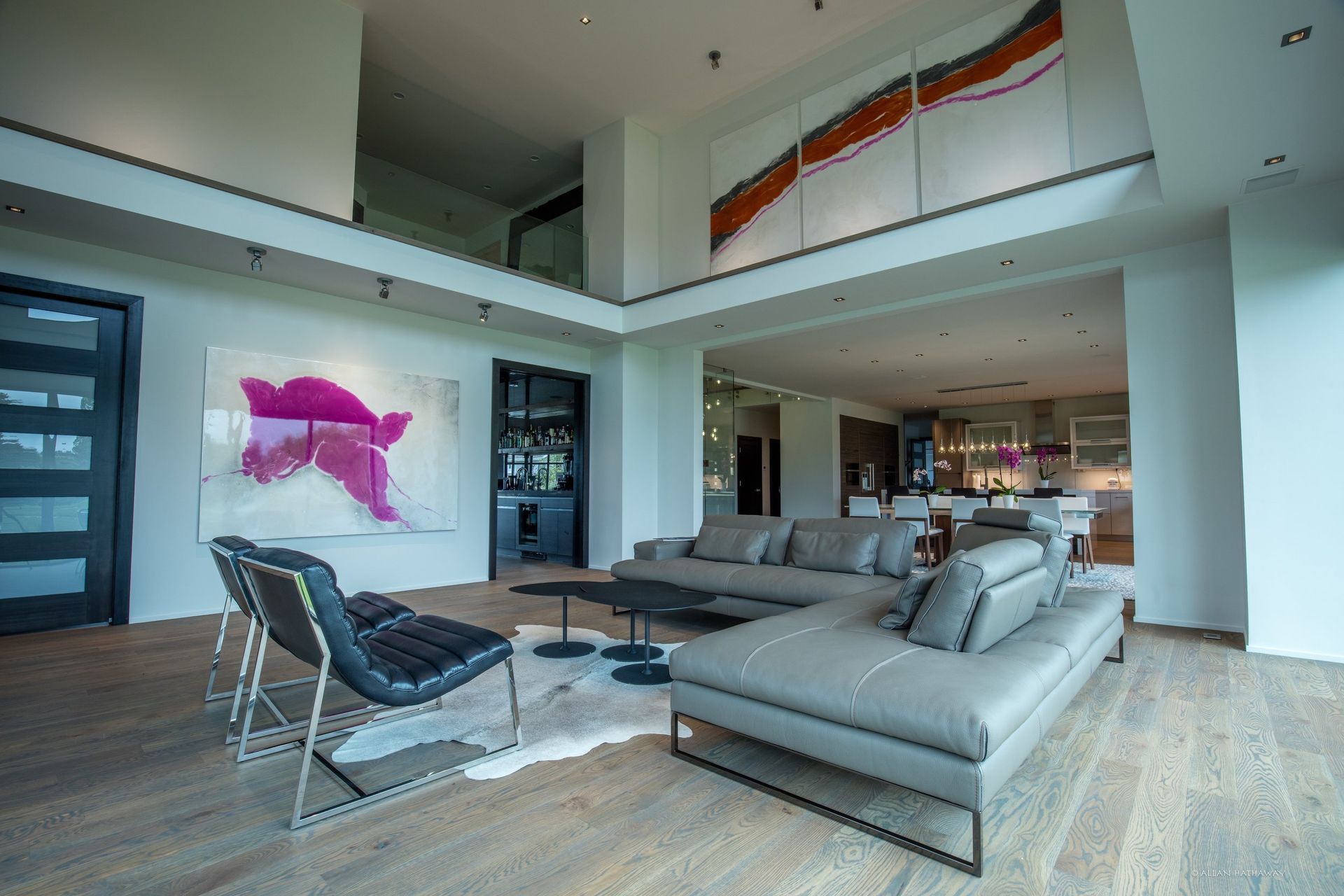 A modern living room with a gray sectional, two black leather chairs, white hardwood floors, and vibrant wall art.