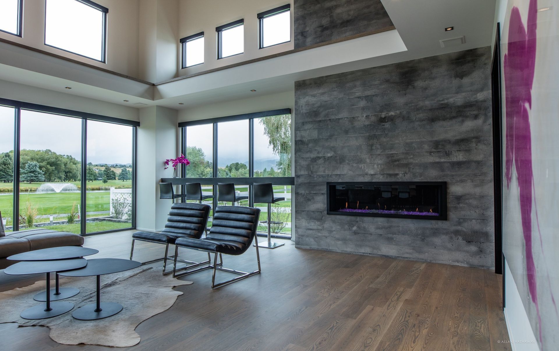 A modern living area with high ceilings, a linear fireplace on a gray stone wall, black chairs, and large window views.
