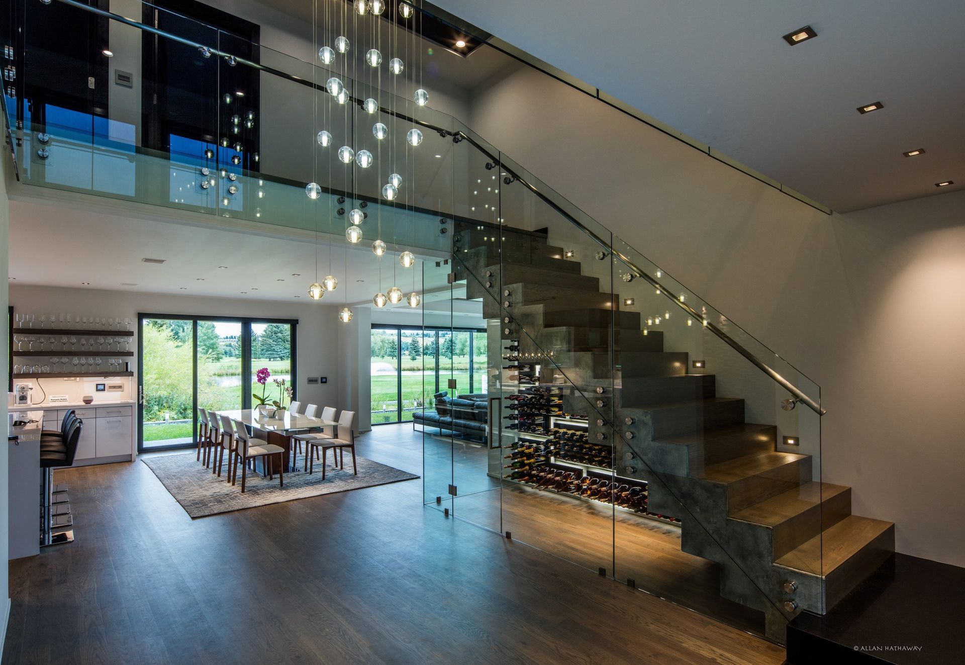 Modern open-plan interior with dark wood floors, a glass-railed staircase, wine storage, and a dining area by large windows.