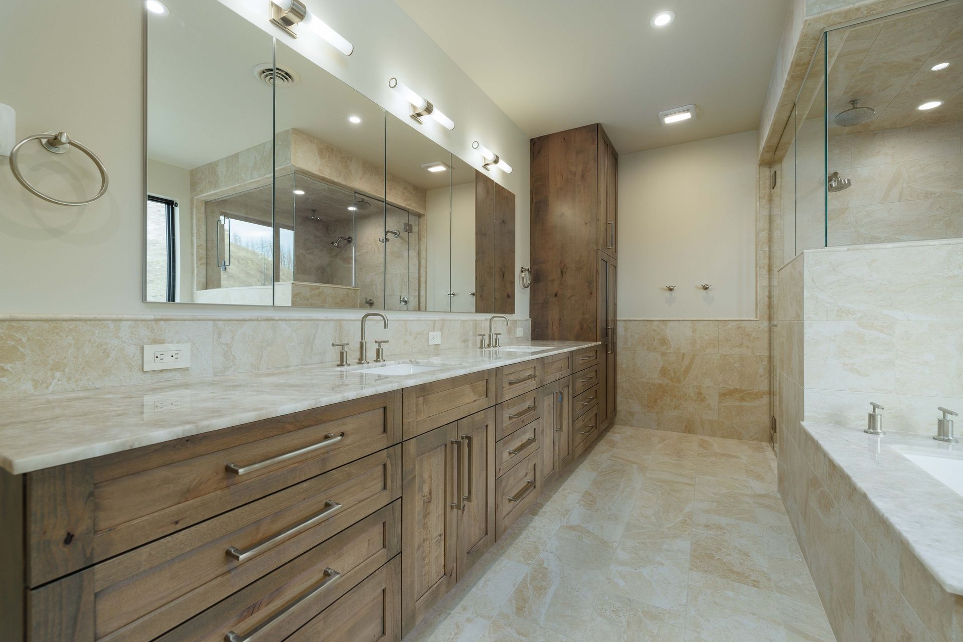 A modern bathroom featuring a long wooden vanity with double sinks, light stone countertops, and a glass-enclosed shower.