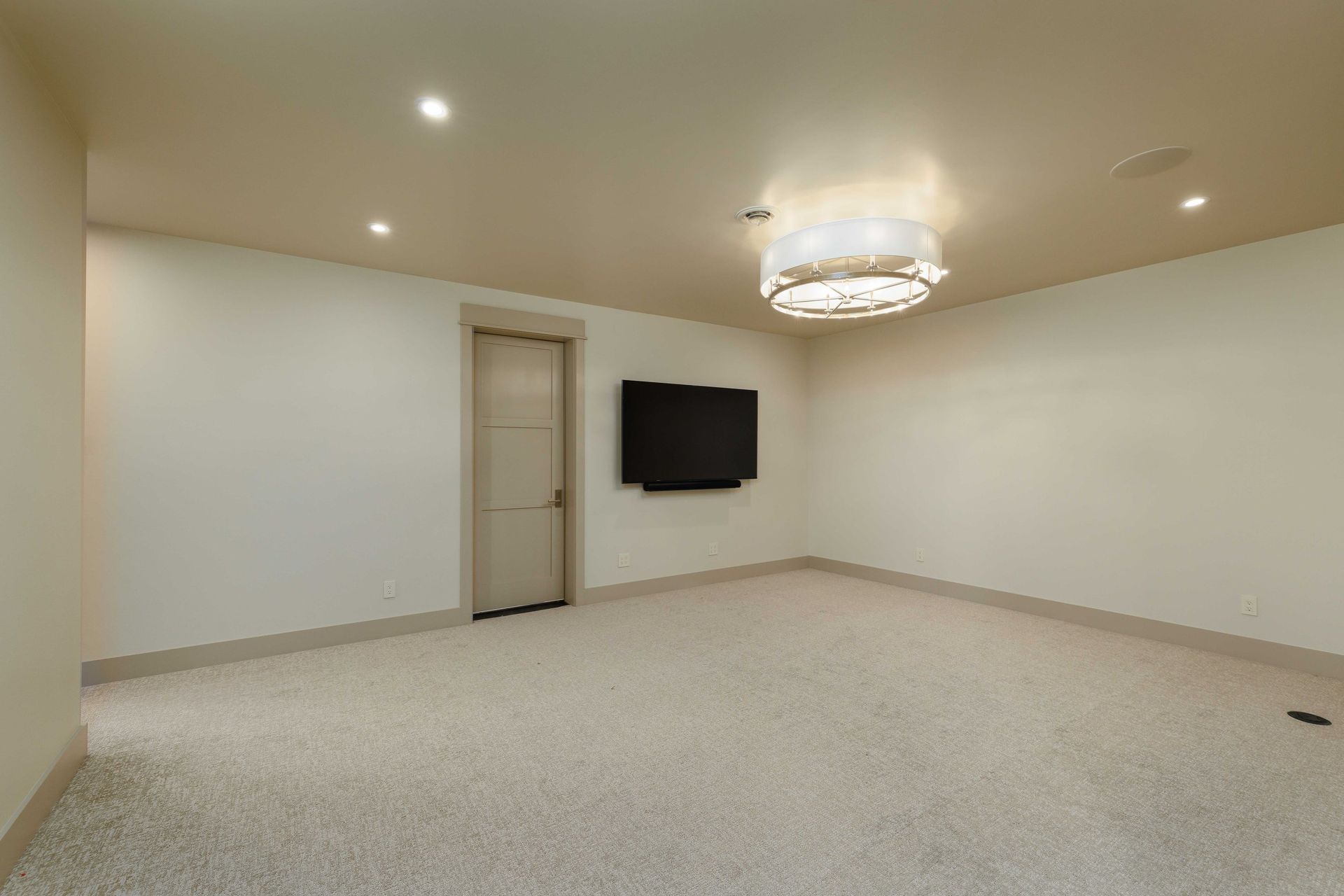A minimalist room with light-colored carpet, plain walls, a closed door, a mounted flat-screen TV, and a drum chandelier.