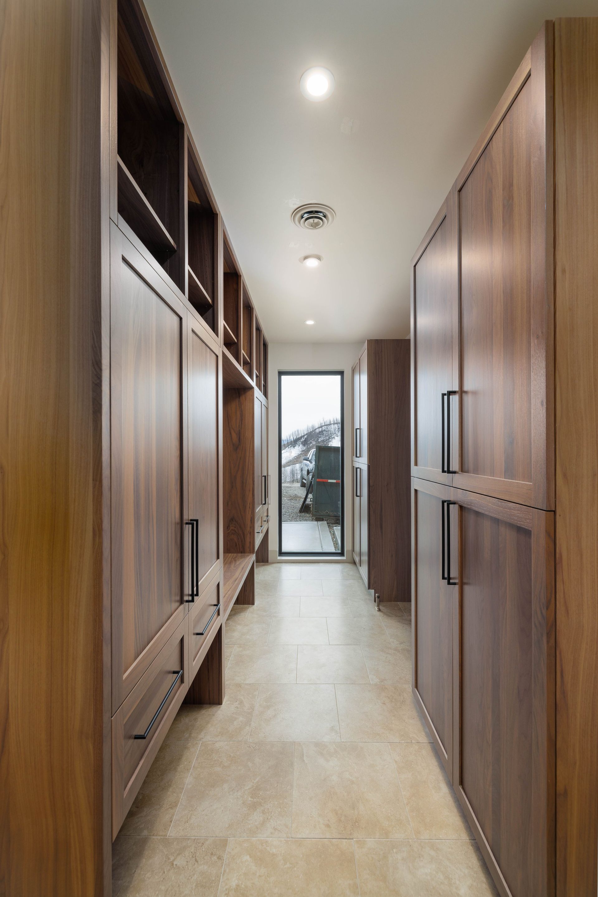 A narrow hallway features floor-to-ceiling wooden storage cabinets on both sides, light tiled flooring, and a glass door.
