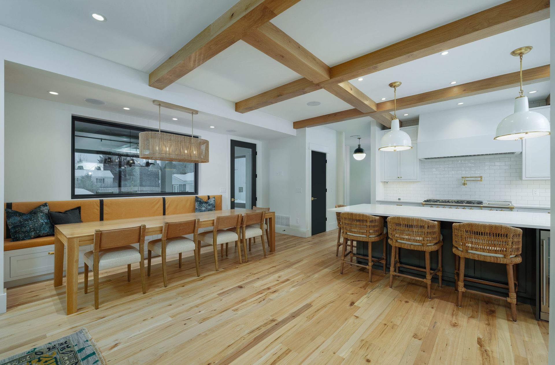 A bright, open-concept kitchen and dining area featuring light wood flooring, exposed wood ceiling beams, and island seating.