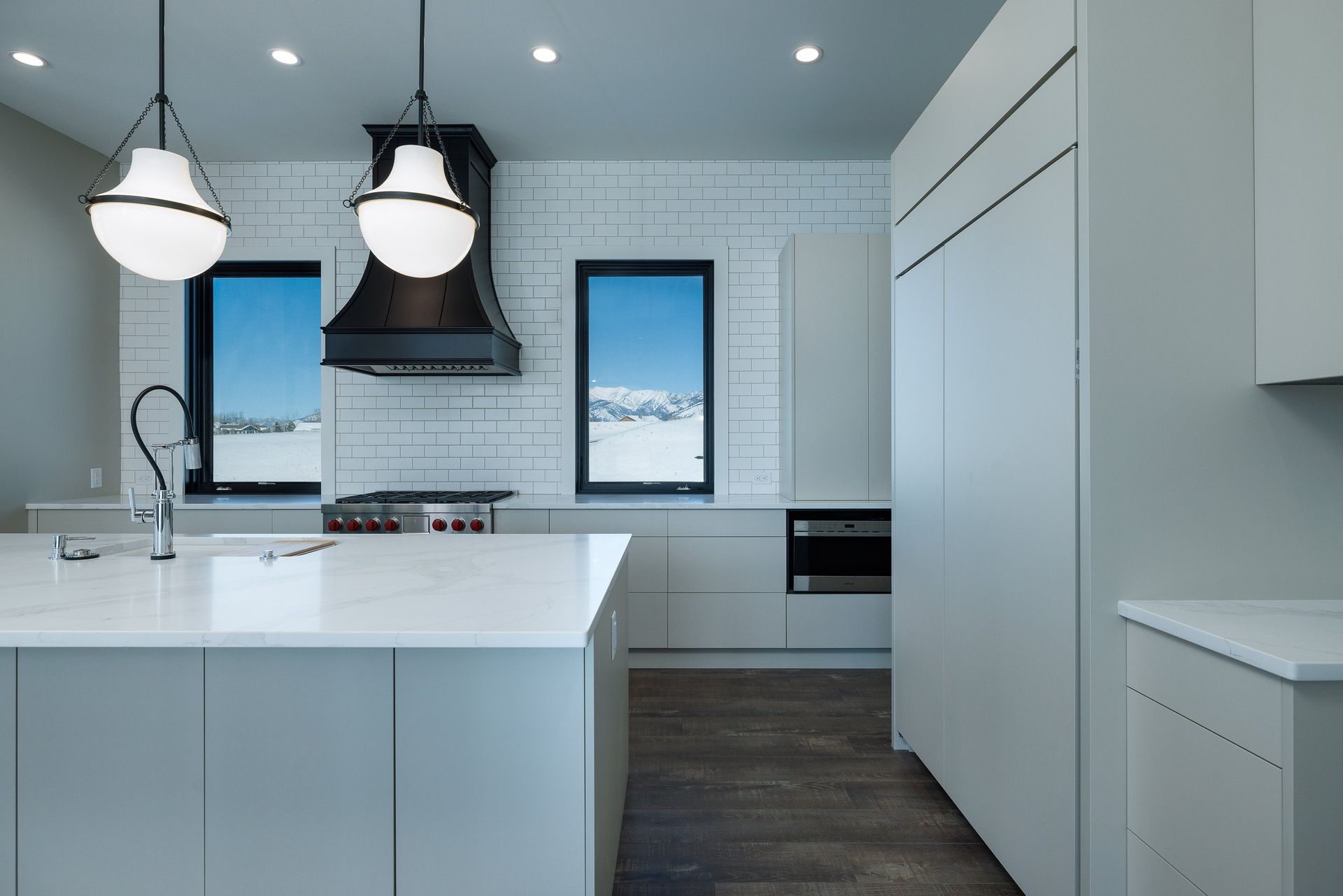 Modern kitchen with a white island, grey cabinetry, two pendant lights, and two windows with snowy outdoor views.