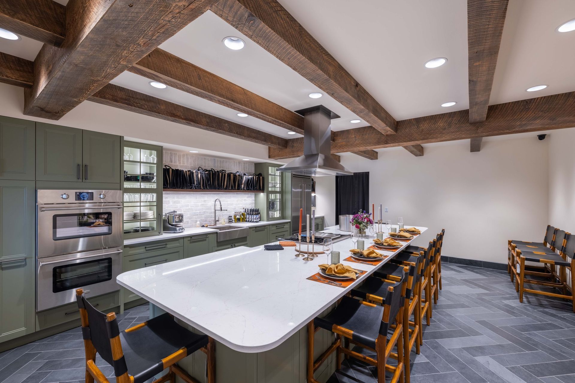 A modern kitchen features sage green cabinets, wood ceiling beams, a large white island with stools, and tiled flooring.