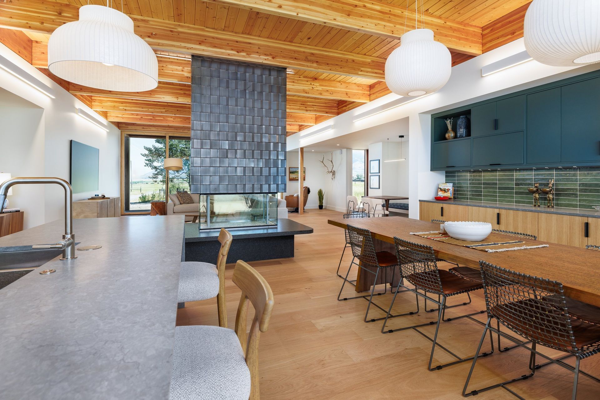 Modern open-plan kitchen and dining area with wood ceiling, stone island, a central fireplace, and views of outdoors.