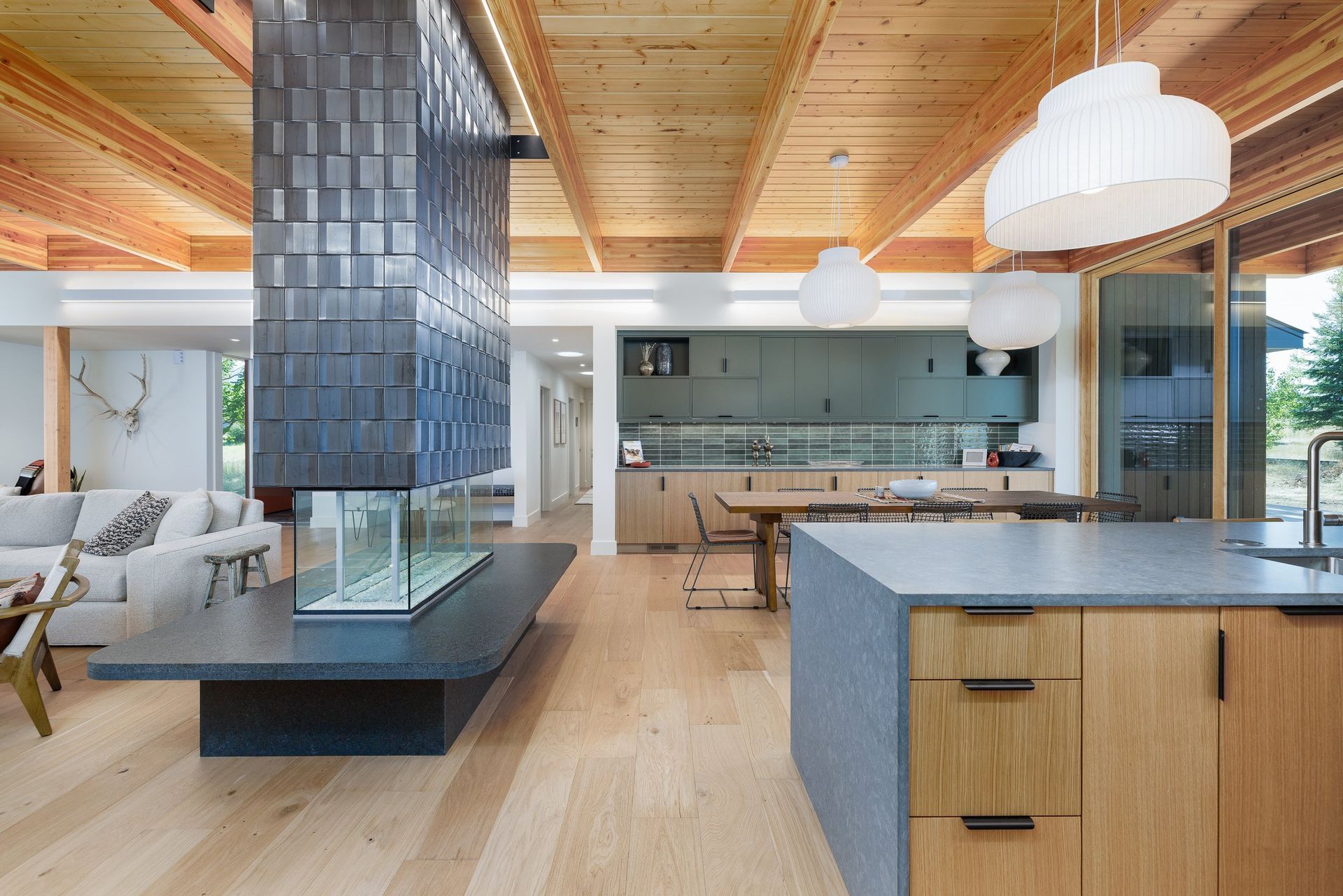 Modern open-concept living area with wood ceilings, a large dark-tiled fireplace, and a kitchen with wooden cabinets.