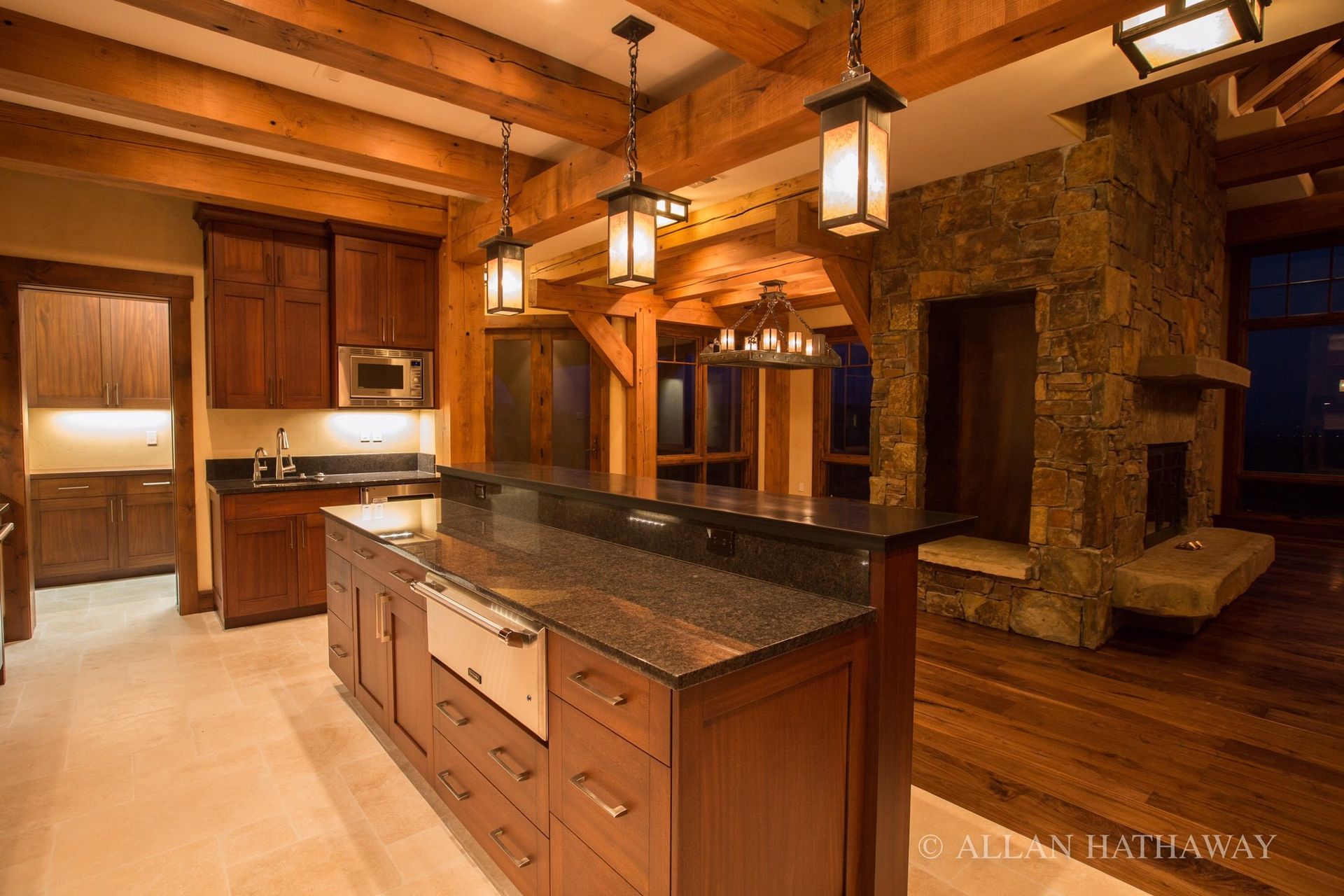 A kitchen with wood cabinets, black granite countertops, stone fireplace, and exposed beams under warm pendant lighting.