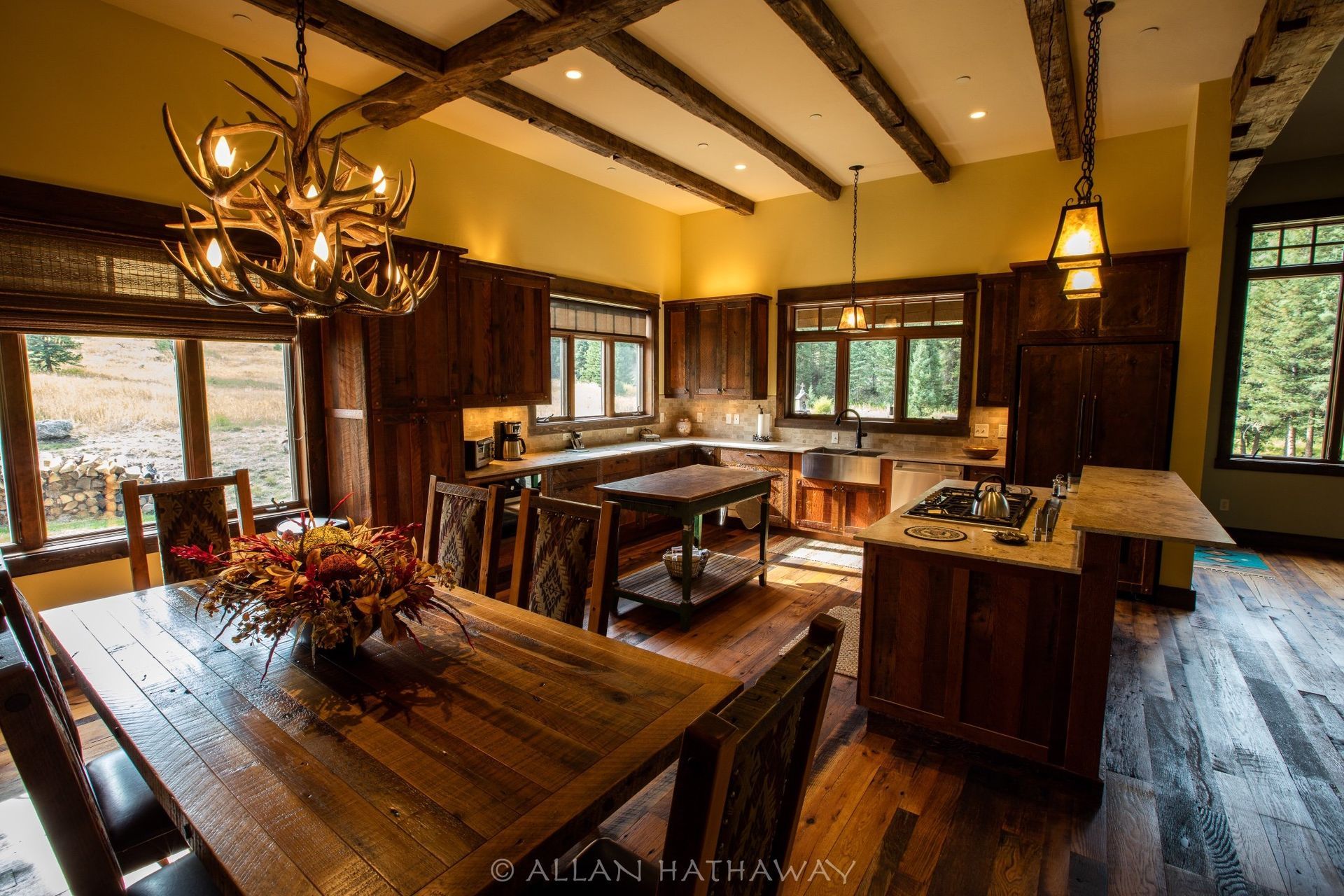 A rustic, open-concept kitchen and dining area with wooden beams, antler chandelier, dark wood cabinetry, and large windows.