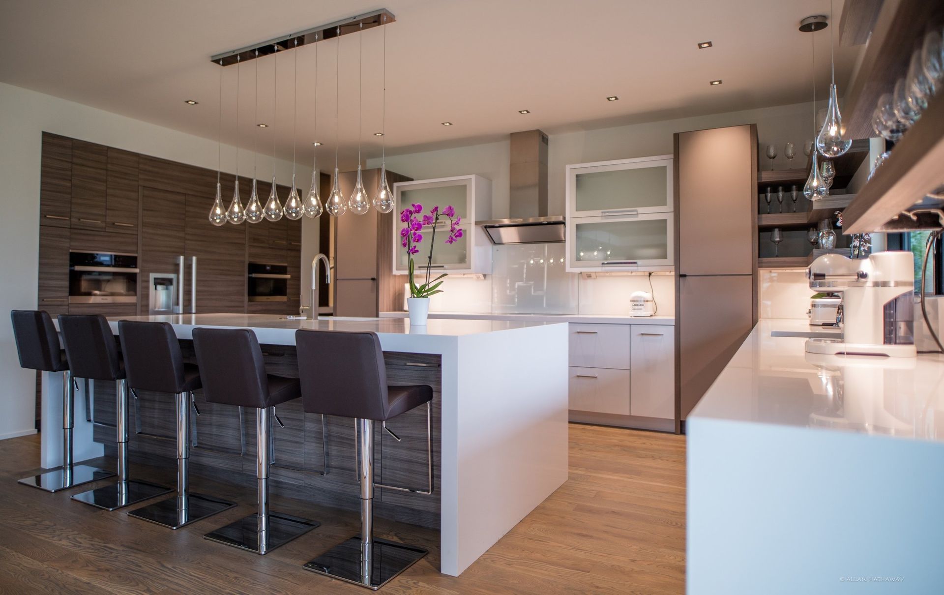 Modern kitchen featuring a white island with four dark stools, pendant lighting, and brown cabinetry.