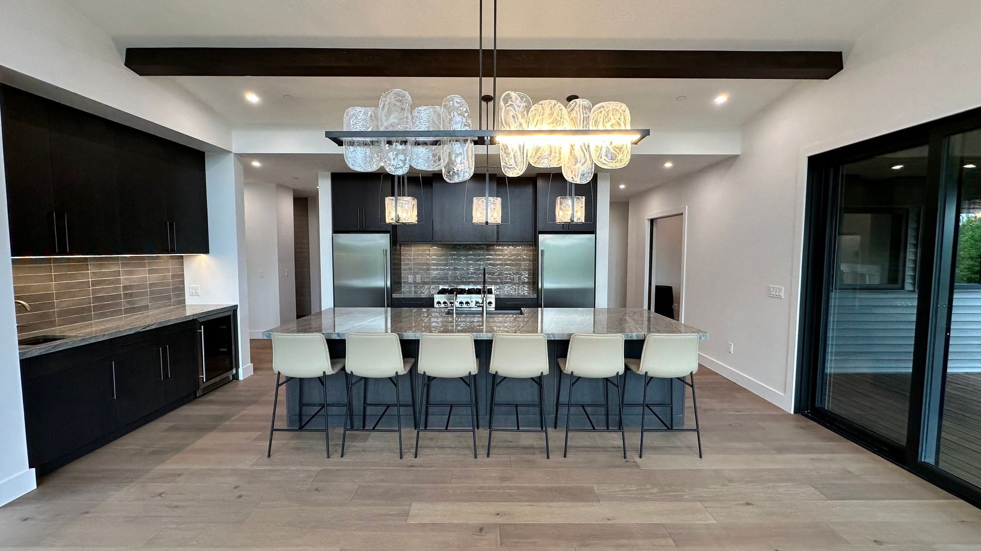 A modern kitchen with dark cabinets, light wood floors, a large central island with six stools, and a glass chandelier.