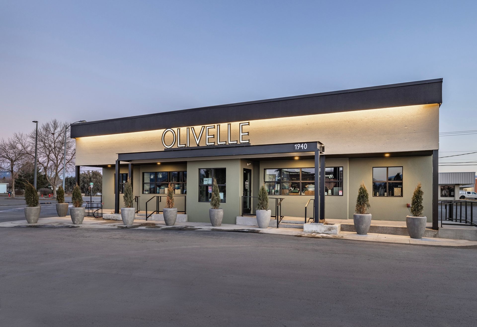 Storefront at dusk with glowing sign, glass windows, and potted plants along the entrance.