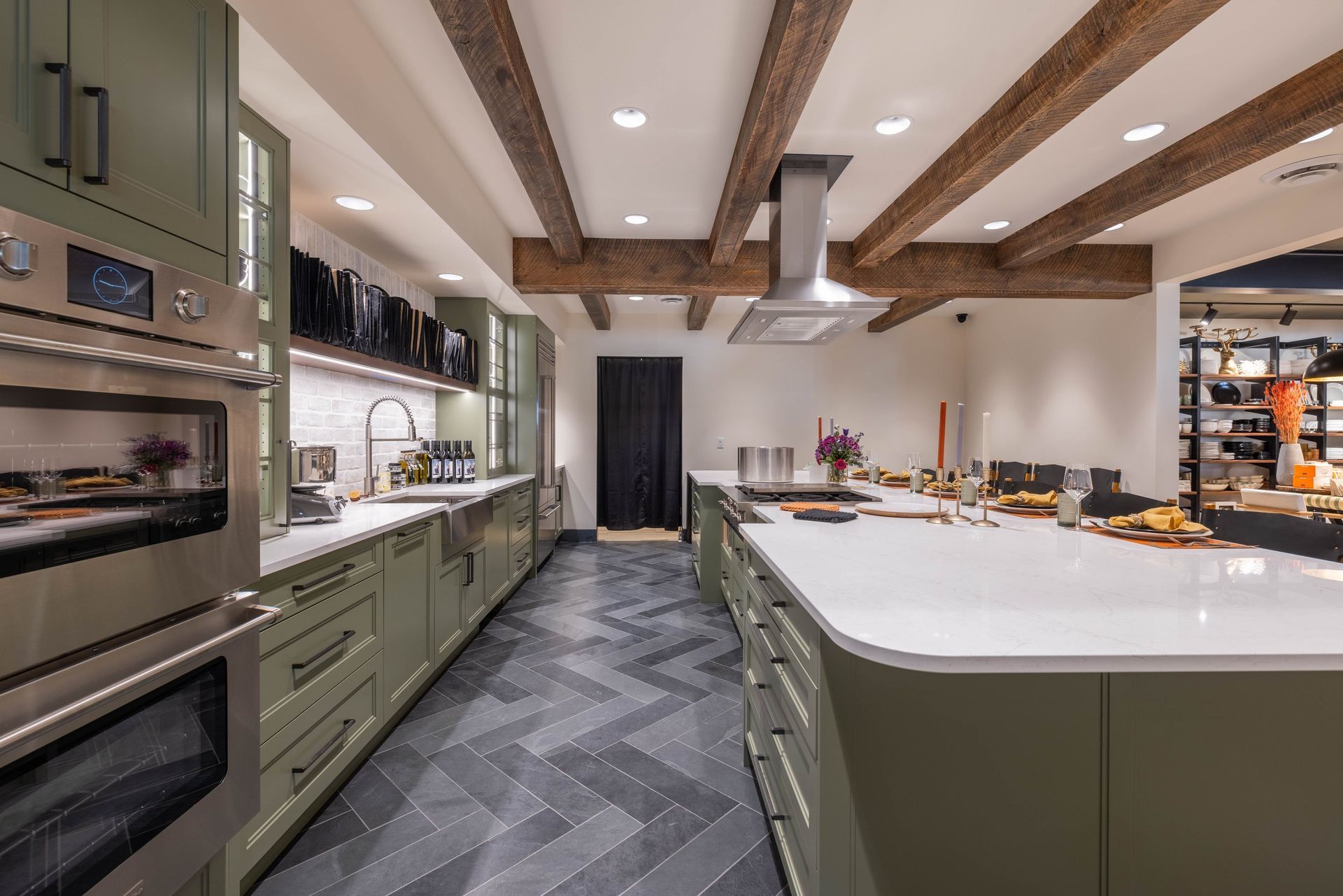 Modern kitchen with sage green cabinets, large white island, dark herringbone tile floor, and exposed ceiling beams.