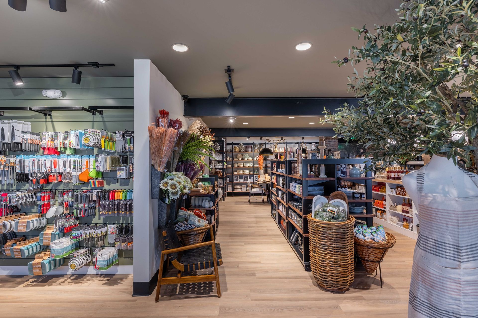 A retail store interior features aisles of kitchenware, a floral arrangement, and woven baskets on light wood flooring.
