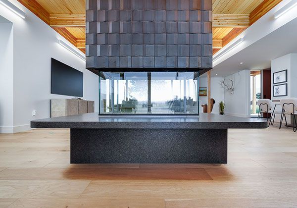 Modern open-concept living room with a large dark fireplace, wood floors, and city views through tall windows.