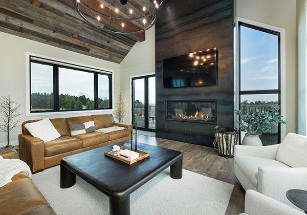 Modern living room with vaulted wood ceiling, fireplace, large windows, tan sofa, and white armchairs.