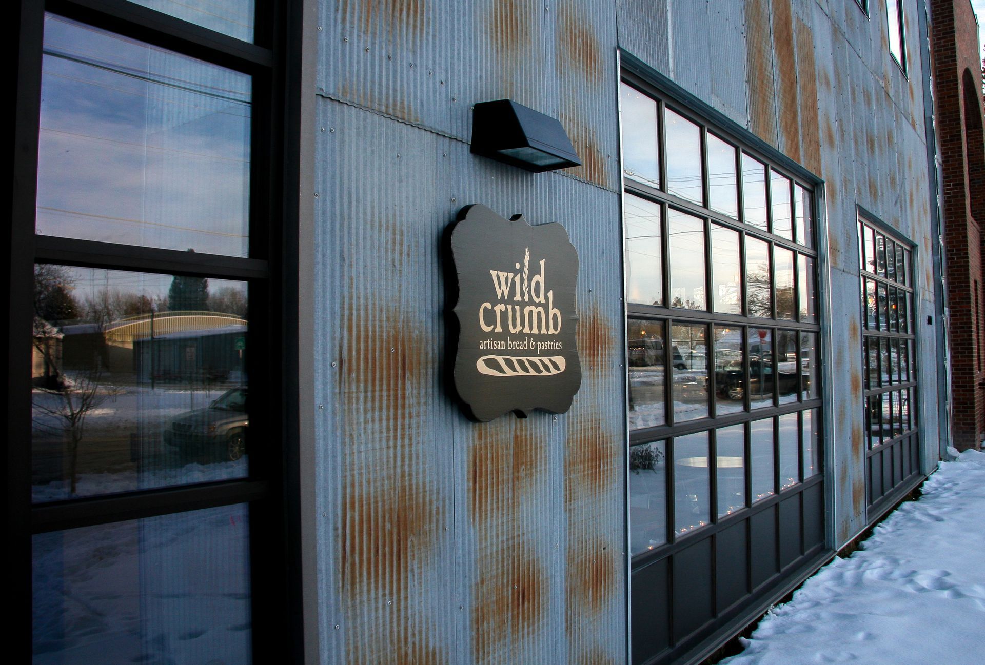 Rustic metal building exterior with a sign and large windows beside a snowy walkway