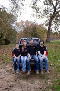 Four people in matching black shirts sit on the lowered tailgate of a pickup truck in a wooded area with fallen leaves. 