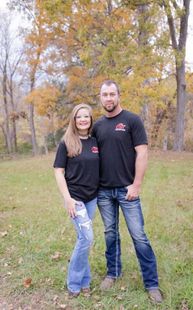 A couple poses together in a field during autumn, wearing matching black t-shirts and jeans in front of fall foliage. 