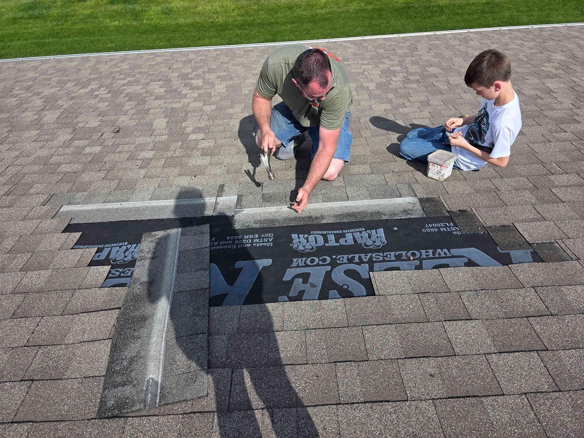 Man and child on a rooftop repairing shingles; man hammers, child assists, sunny day.