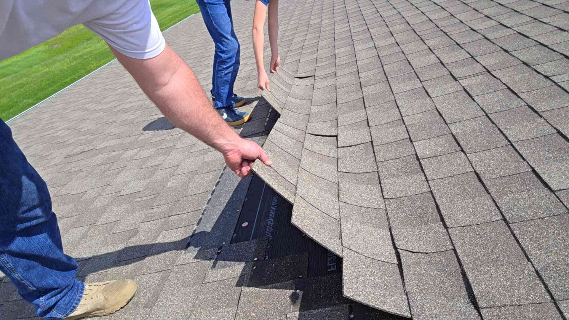 Two people examine a damaged asphalt shingle roof.