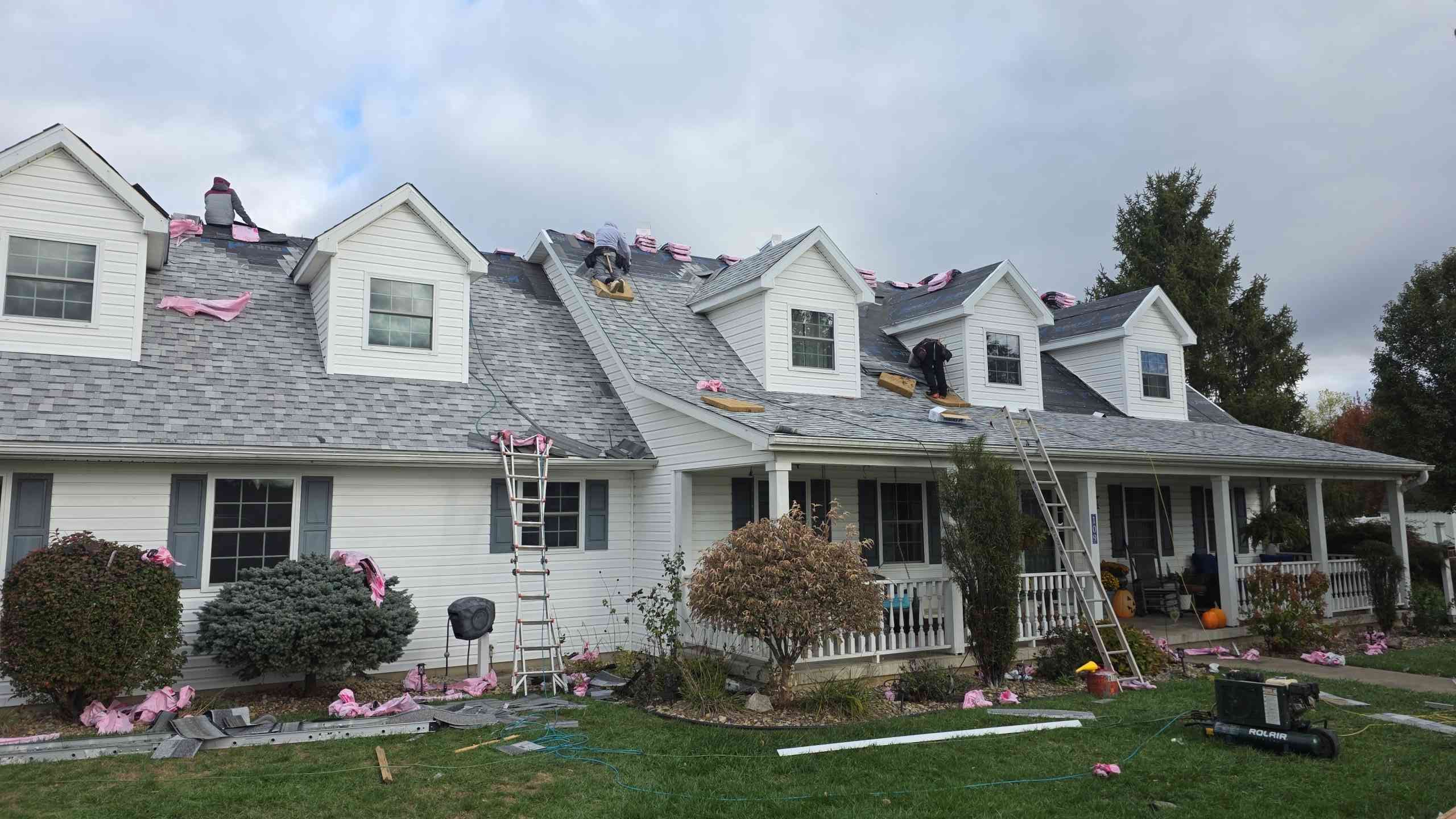 Roofers replacing shingles on a white house with multiple dormers under a cloudy sky.