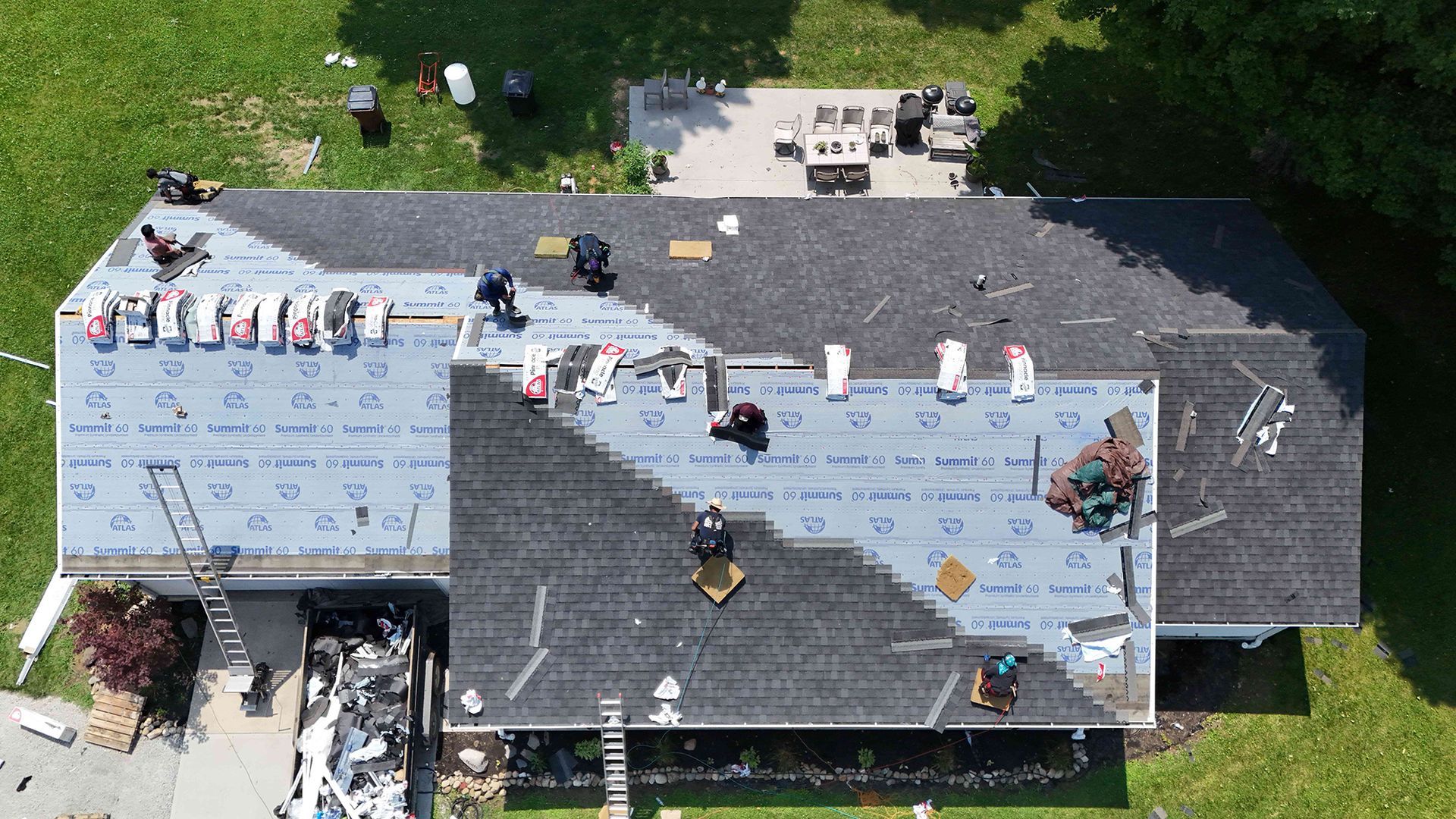 Roofers working on a house roof from an aerial view. Shingles, materials, and workers are visible.