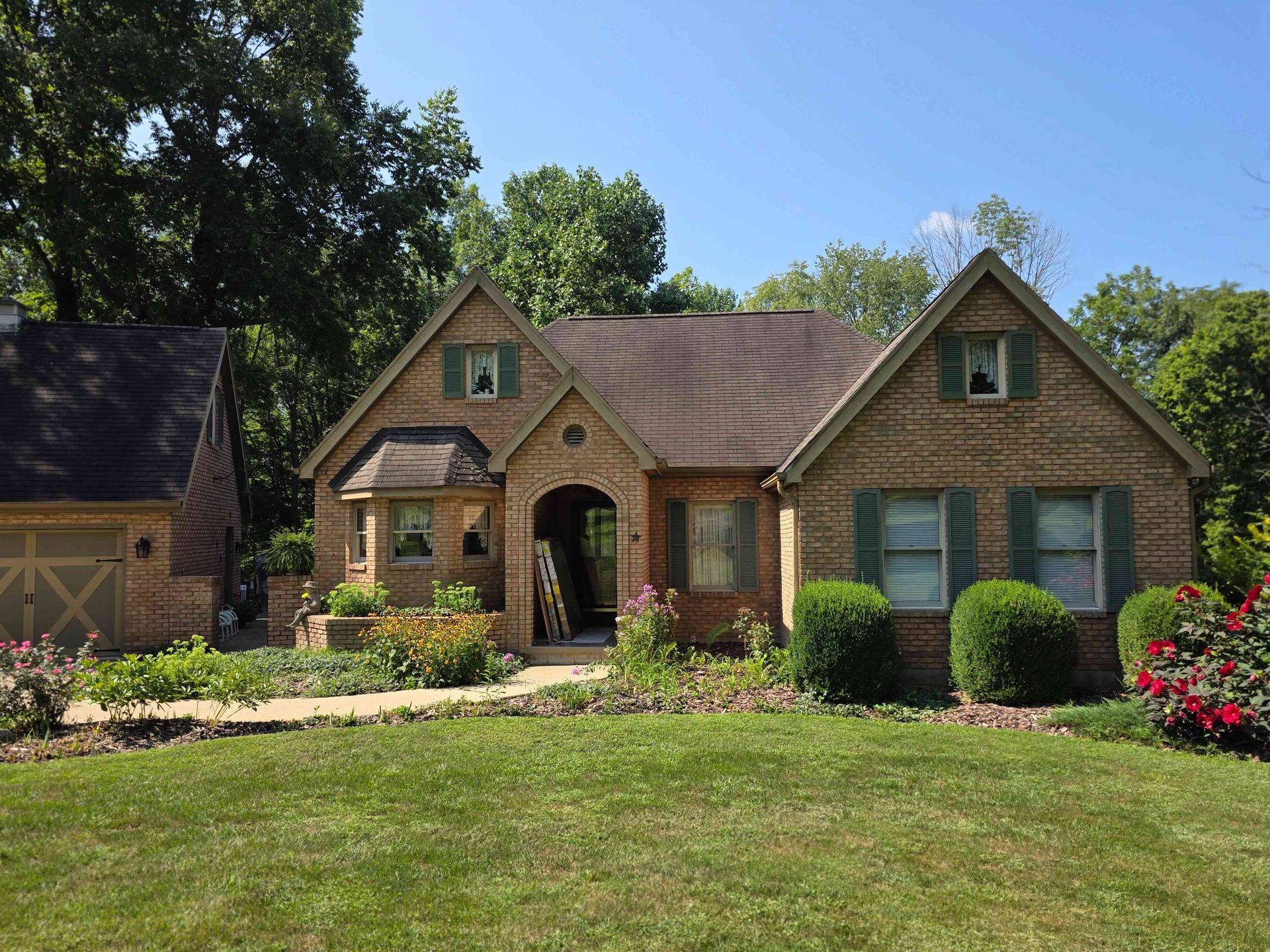 Brick house with green shutters and brown roof; flowers in the front yard.