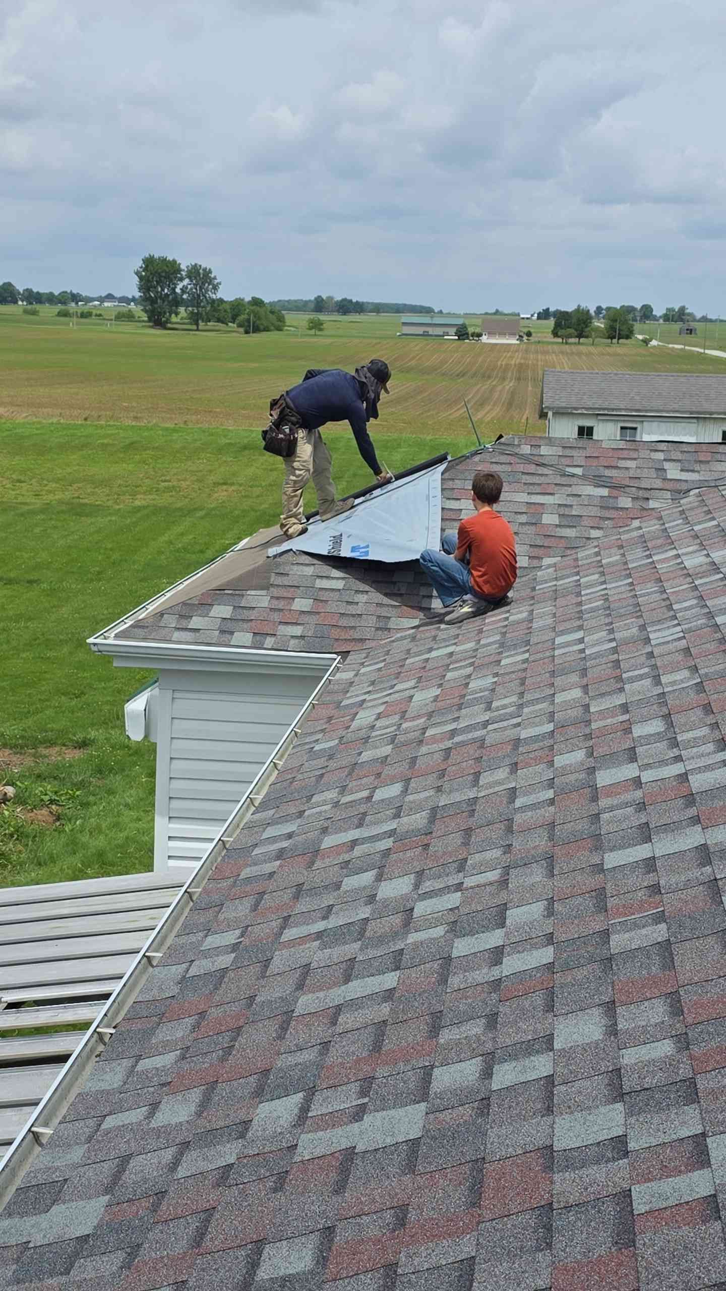 Two people working on a roof, one holding a piece of metal, against a rural backdrop.