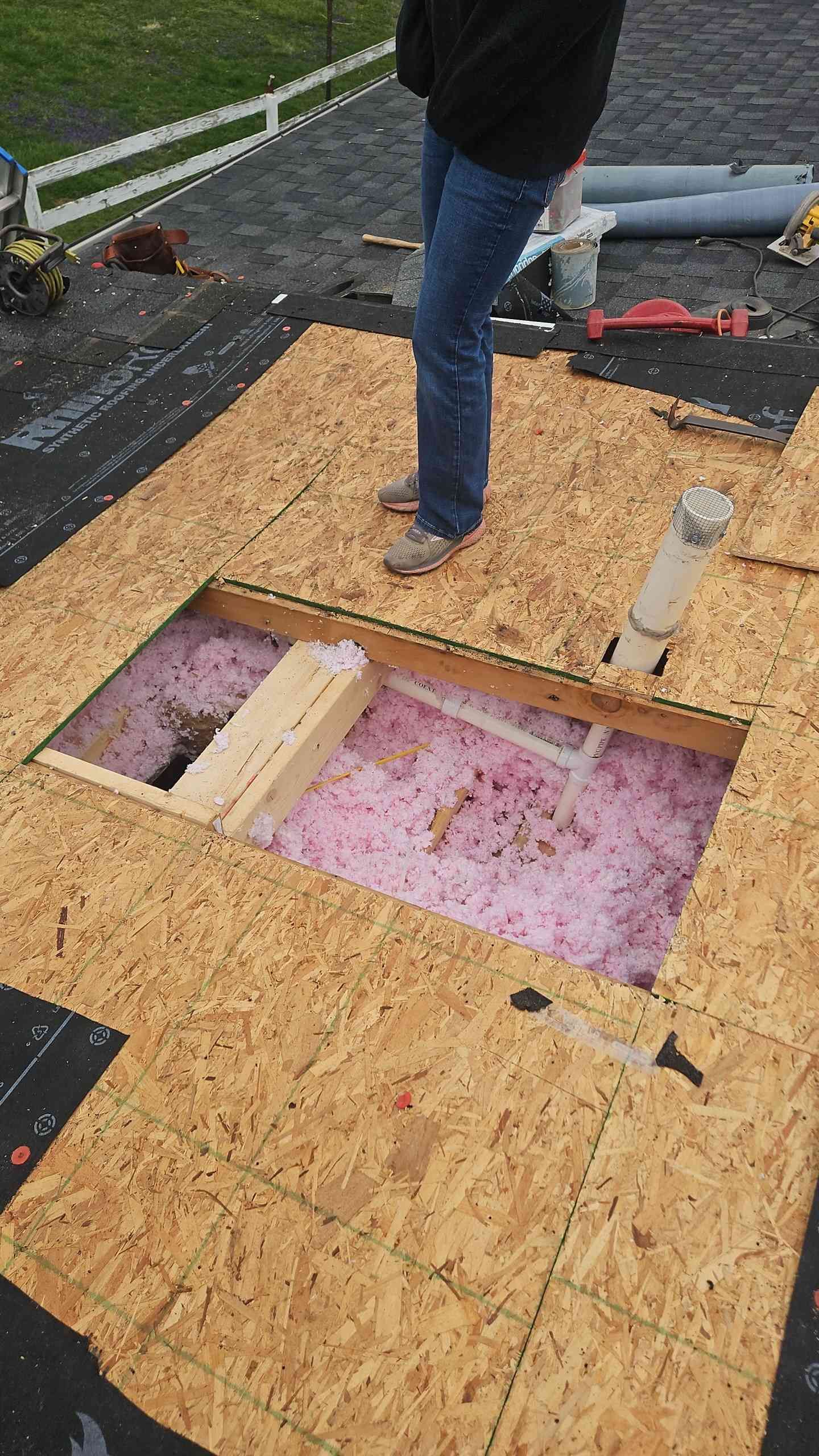 Person on a rooftop works on insulation around a rectangular opening in the wood, pink insulation visible.