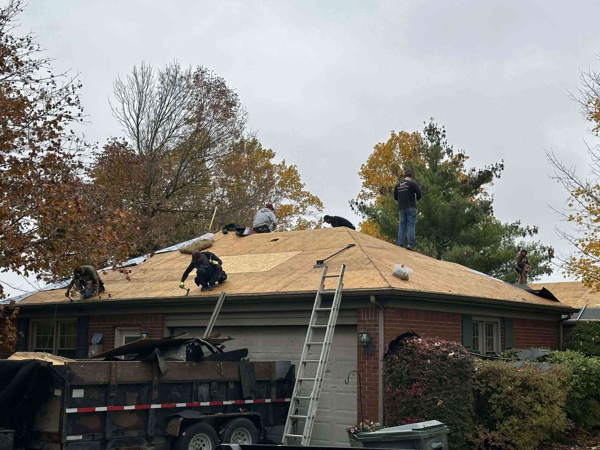Roofers installing shingles on a house with a red brick exterior, trailer, and trees in the background.