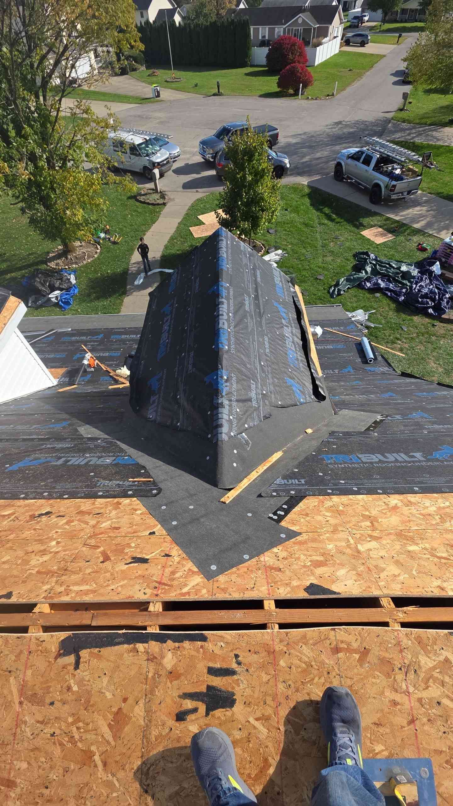 Roofer on a partially shingled roof, looking down.  Roofing materials are visible. Suburban neighborhood in the background.